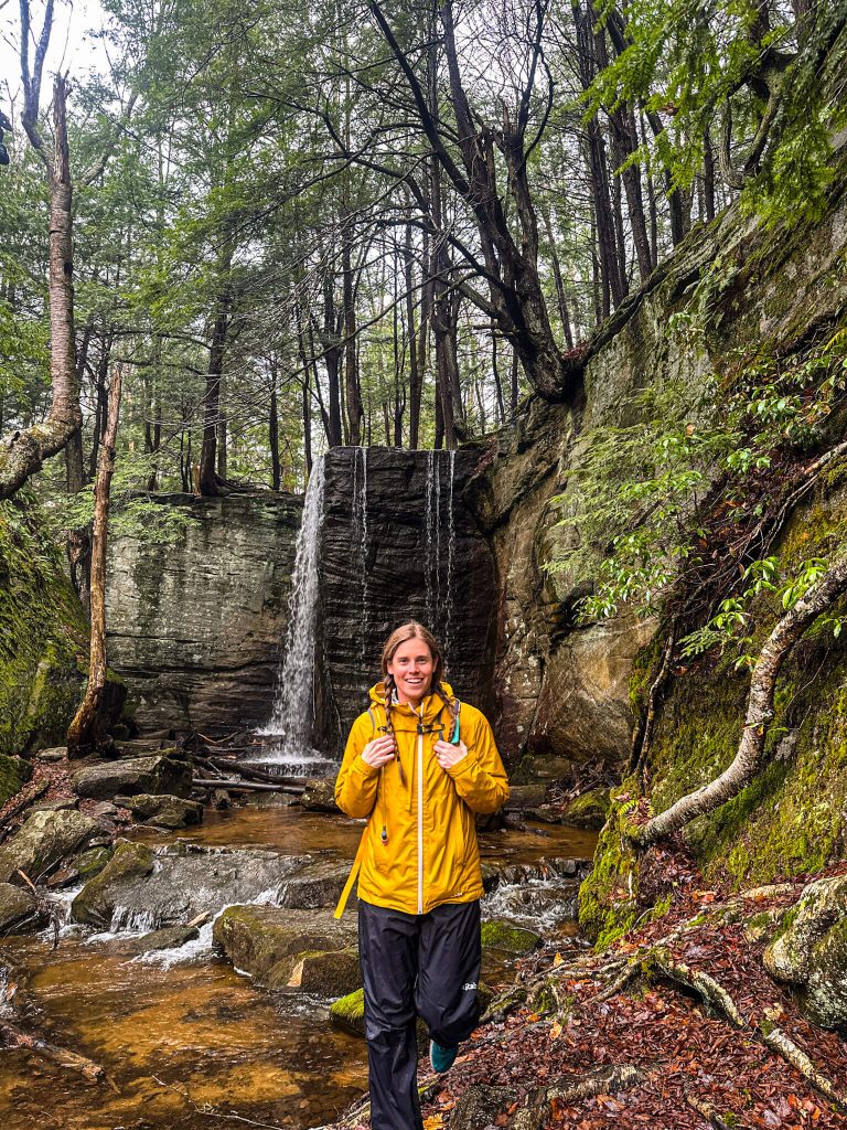 A hiker in a bright yellow jacket stands in a shallow stream beneath a small waterfall surrounded by moss covered rock walls and dense forest.