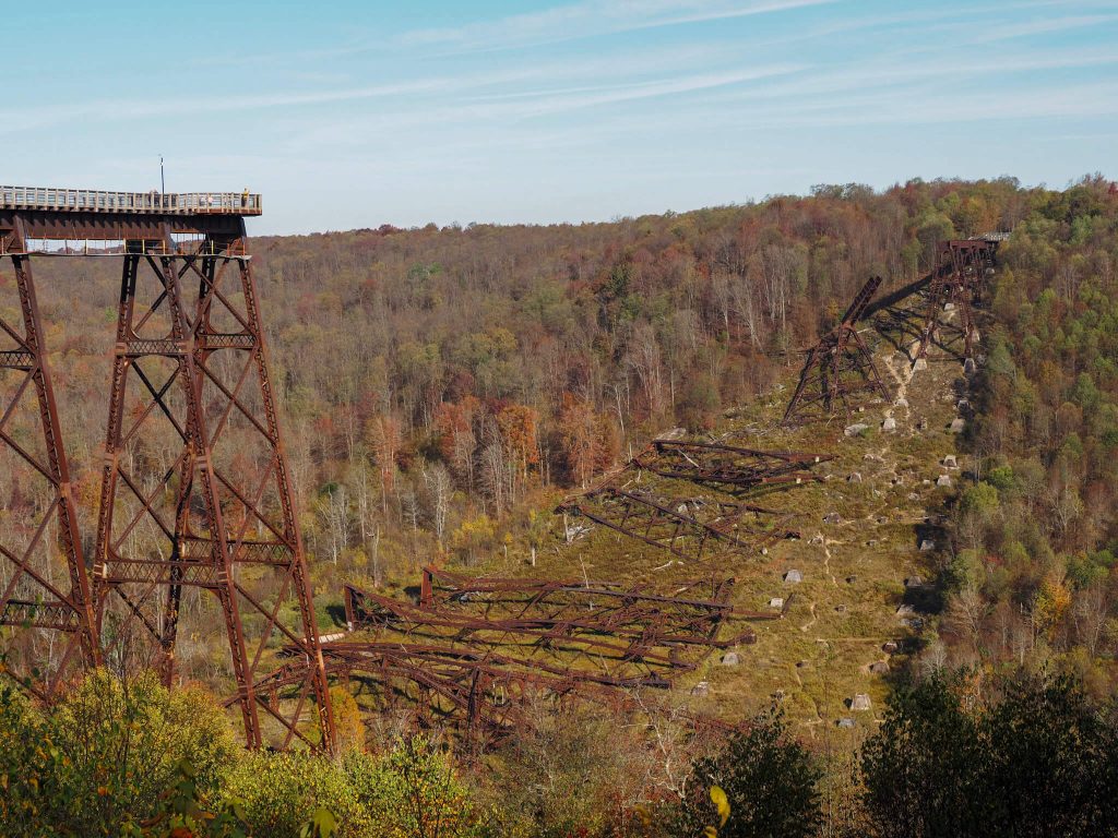 Kinzua Bridge State Park in autumn, showing the remains of the historic Kinzua Viaduct scattered across a grassy valley beneath a bright blue sky. Rusted steel towers and twisted bridge sections stand among colorful fall trees and vegetation.